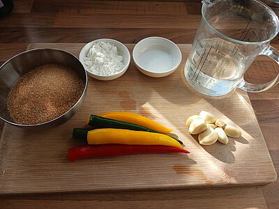 Ein Schneidbrett mit einer Schüssel braunem Zucker, Chilies, Knoblauch, einem Messbecher sowie zwei Schälchen mit Salz und Stärke